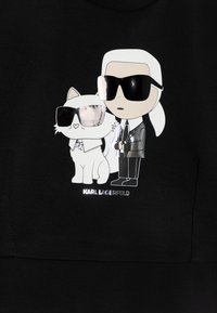 Melns kapucis džemperis ar karikatūru varoņa attēlu, kur vīrietis uzvalkā un baltais kaķis, abi nēsā saulesbrilles; zem attēla ir ieraksts "KARL LAGERFELD".
