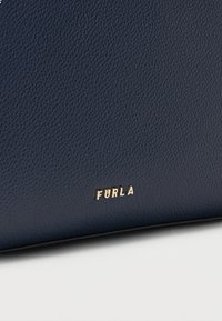 Teksturirana usnjena površina mornarsko modre barve z majhnimi zlatimi kovinskimi črkami, ki na spodnjem robu tvorijo besedo "FURLA".