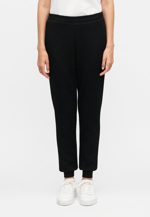 Personne portant un pantalon de jogging noir resserré aux chevilles avec revers et des baskets blanches, debout devant un fond blanc uni.