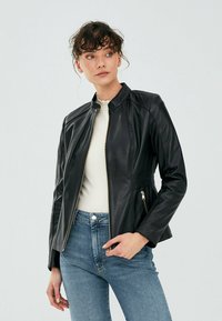 Giubbotto di pelle nera con colletto alto, design aderente e tasche con zip. La texture liscia e i dettagli delle cuciture sottili esaltano il look.