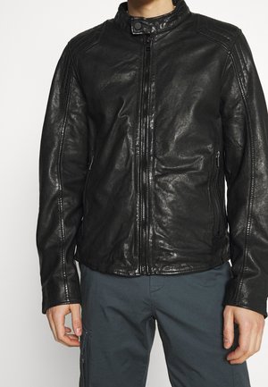 Homme portant une veste en cuir noire zippée avec un col pression et un pantalon gris foncé sur un fond clair.