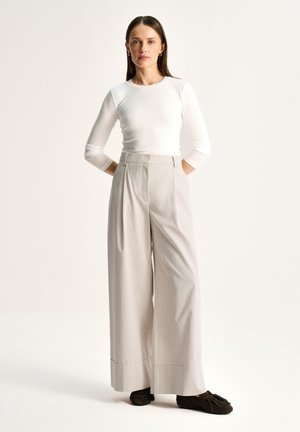 Kobieta stojąca z rękami z tyłu, ubrana w biały top z długim rękawem, szerokie beżowe spodnie z wysokim stanem i ciemne mokasyny na jasnym tle.