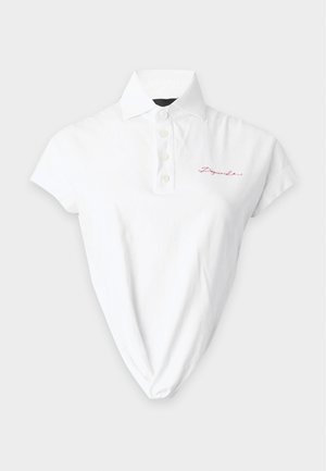 Jeune femme aux longs cheveux foncés porte un polo blanc à manches courtes rentré dans un pantalon gris ample avec une ceinture rouge, debout les mains dans les poches.
