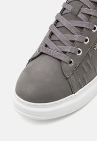 Sivi kožni tenisice s ravnim vezicama, metalnim rupicama i bijelim gumenim potplatom; imaju teksturirani bočni uzorak i glatki vrh.