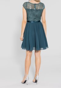 Robe teal avec un bodice pailleté et une jupe fluide. Le design supérieur présente des manches courtes. Portée avec des talons argentés.