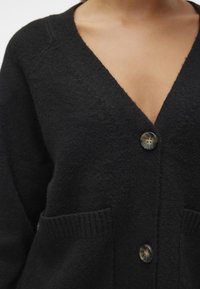 Cardigan noir avec un col en V, deux poches devant, fermeture à boutons, poignets côtelés et tissu doux et texturé.