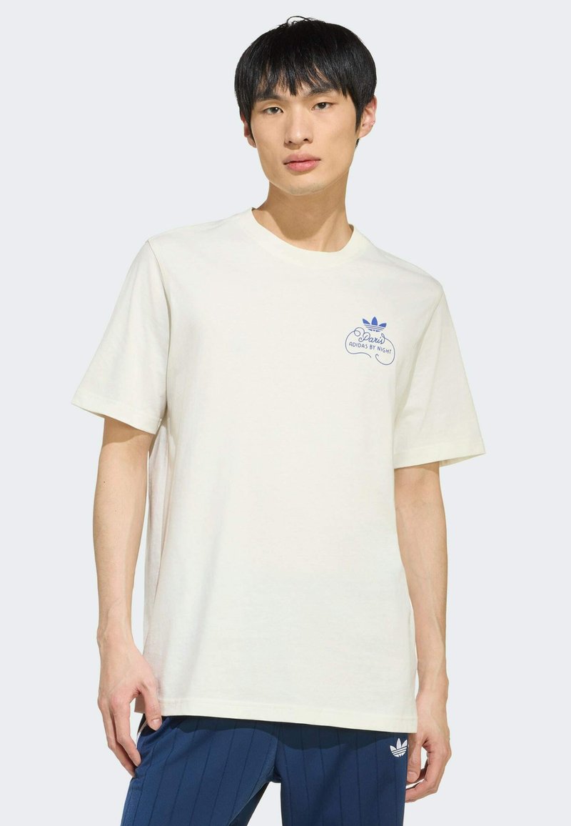 Jeune homme portant un t-shirt blanc Adidas avec un petit logo bleu et le texte « Paris Adidas by Night », assorti à un pantalon Adidas bleu.