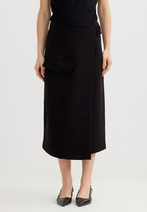 Mujer con falda midi cruzada negra con un gran bolsillo en el lado izquierdo y un detalle de hebilla en la cintura, combinada con tacones negros puntiagudos.