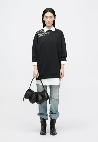 Czarna, oversize, sweatshirt z haftowanymi akcentami, nałożona na białą koszulę. W połączeniu z jasnoniebieskimi dżinsami i czarną skórzaną torbą.