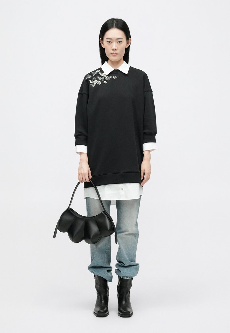 Zwarte oversized sweatshirt met geborduurde accenten, gelaagd over een wit shirt. Gecombineerd met lichtblauwe jeans en een zwarte leren tas.