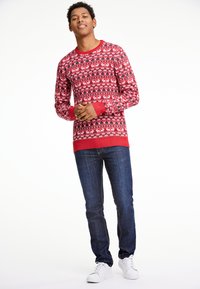 Röd stickad tröja med ett julmönster av vita renar och snöflingor, kombinerad med mörkblå jeans och vita sneakers.