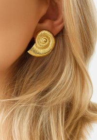 Orecchino a spirale dorato con texture rigata, che imita un guscio di nautilus, posizionato contro capelli biondi.