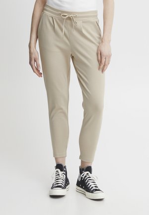 Beige joggebukser med elastisk liv, snor og smale anklonstrikk, kombinert med svarte høye sneakers.