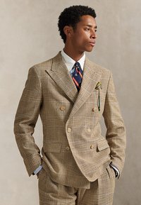 Uomo che indossa un completo doppiopetto a quadri beige e marroni con camicia bianca, cravatta a righe blu navy e fazzoletto giallo nel taschino, con le mani in tasca.