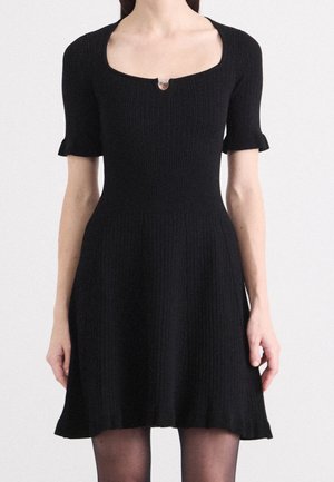 Woman wearing a fitted black ribbed knit dress with short sleeves and a small silver clasp at a square neckline.