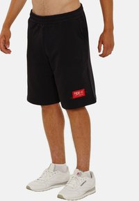 Schwarze Sportshorts aus weichem Material, mit elastischem Bund und seitlichen Taschen, ausgestattet mit einem roten Logopatch am unteren Bein.