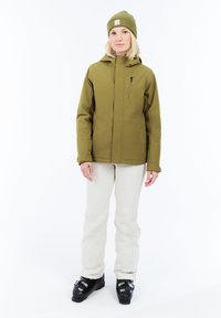 Žena stojí oblečená v olivovo zelenej bunde a čiapke, bielych nohaviciach a čiernych lyžiarskych topánkach na bielom pozadí.