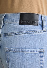 Gaišzilas džinsi ar melnu ādas DKNY uzlīmi jostasvietā. Iespējo aizmugurējās kabatas un izbalējusi tekstūra. Vienkārša šuvju detalizācija.