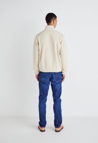 Chaqueta de forro polar beige con un cuello alto, combinada con jeans de mezclilla azules. La chaqueta tiene puños acanalados y un corte recto. Zapatos con acentos naranjas.