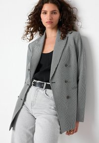Grijze geblokte blazer met dubbele knopen, met een gesneden revers en textielstof, gecombineerd met een zwarte top en lichtgrijze jeans.