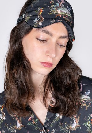 Black sleep mask with floral and bird patterns, featuring a soft fabric and curved shape, worn by a person with brown hair.