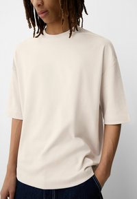 Világos bézs színű, rövid ujjú t-shirt puha anyagból, laza szabású, kerek nyakkivágással és minimális varrásokkal, sötét farmernadrággal párosítva.