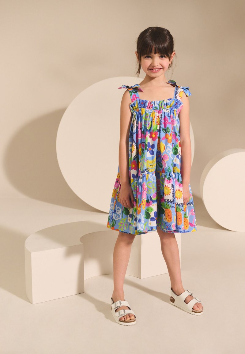 Vestito floreale colorato con base blu, scollo arricciato e spalline annodate. Abbinato a sandali bianchi, in piedi su forme di sfondo neutro.