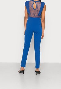 Kungablå overall med volangkrage, genomskinlig spetsbak, och välanpassade ben. Har ett nyckelhålsdetalj vid nacken och en mjuk textur.