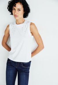 Hvit ermeløs topp med utsmykkede krystall detaljer på skuldrene, kombinert med mørk blå jeans, som viser en figursydd silhuett.