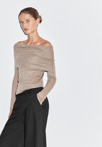 Top color marrone chiaro con scollo a spalle scoperte e maniche lunghe, caratterizzato da un design drappeggiato. Abbinato a pantaloni neri a vita alta e gamba larga.