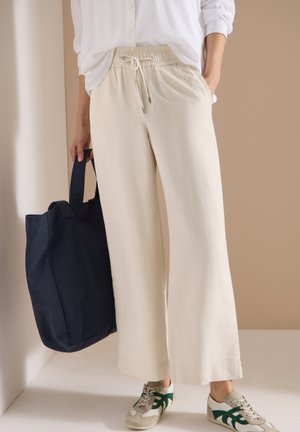 Personne portant un pantalon beige ample avec cordon, une chemise blanche, des baskets beige et vert, tenant un grand sac fourre-tout bleu foncé, debout à l'intérieur.