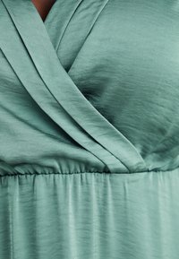 Turkoosi mekko pehmeästä, satiinimaisesta kankaasta, jossa on kääritty vartalon muoto. Vyötärö on kiristetty elastisella vyöllä, luoden pehmeitä laskoksia.