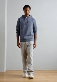 Kapuzenpullover in schieferblau, weicher Stoff mit einer Fronttasche. Kombiniert mit hellgrauen Jeans und weißen Sneakers auf einem Holzboden.