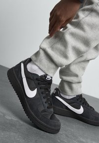 Baskets en suède noires avec le swoosh blanc de Nike, tissu texturé, bout rond, lacets plats et semelles en caoutchouc, portées avec un pantalon de survêtement gris.