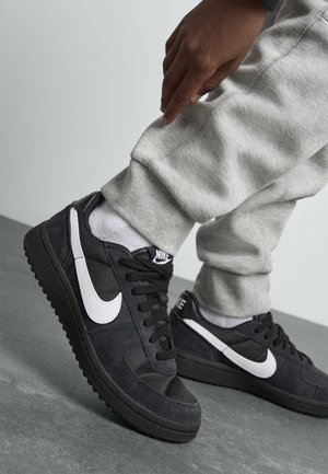 Baskets en suède noires avec le swoosh blanc de Nike, tissu texturé, bout rond, lacets plats et semelles en caoutchouc, portées avec un pantalon de survêtement gris.