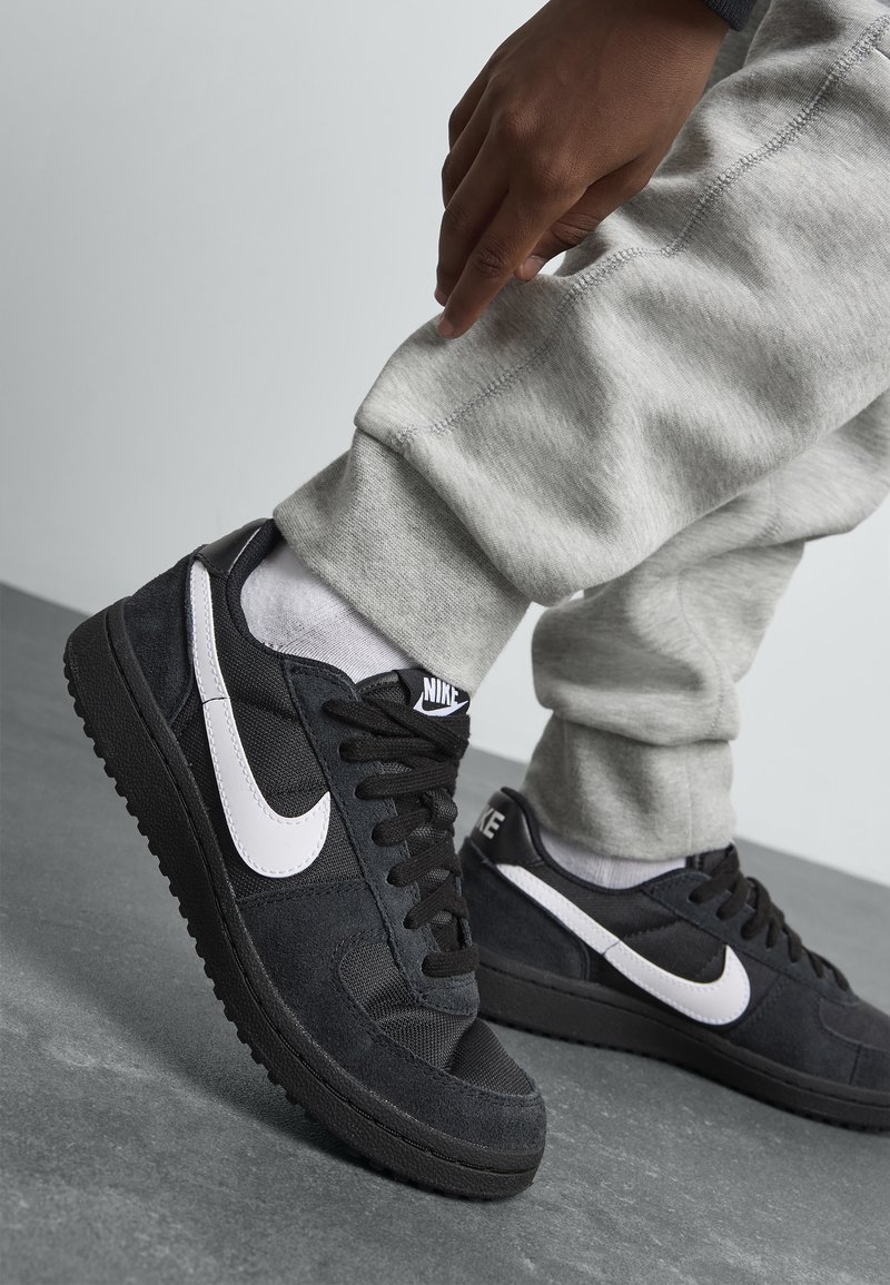 Baskets en suède noires avec le swoosh blanc de Nike, tissu texturé, bout rond, lacets plats et semelles en caoutchouc, portées avec un pantalon de survêtement gris.
