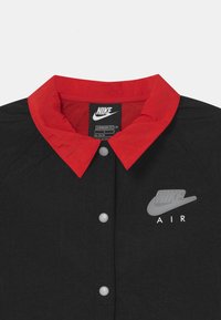Černá bunda s červeným límcem, vyrobená z texturované látky a s bílými puklicovými knoflíky. Obsahuje šedé logo Nike a text "AIR".