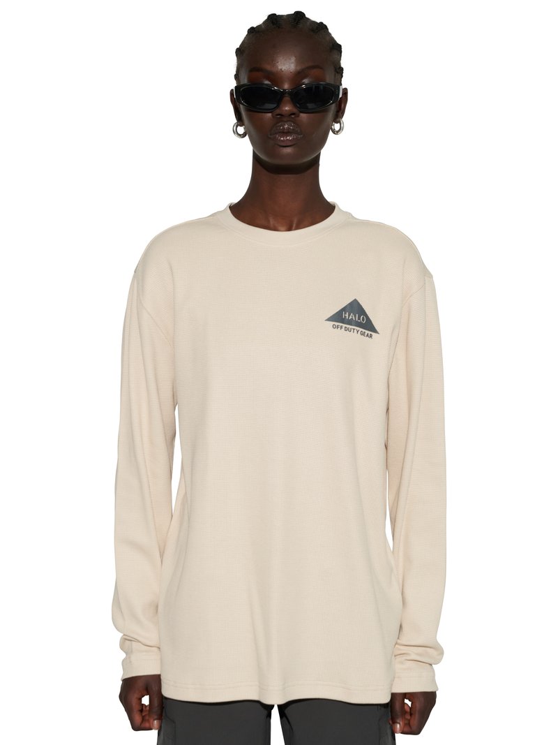 Osoba ubrana w kremową koszulę z długim rękawem z logo "HALO OFF DUTY GEAR", czarne okulary przeciwsłoneczne, srebrne kolczyki-koła i ciemne spodnie, stojąca przodem.