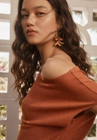 Pull rouille à épaules dénudées avec un tricot texturé. Modèle portant des boucles d'oreilles florales, ayant de longs cheveux ondulés et un fond légèrement surexposé.