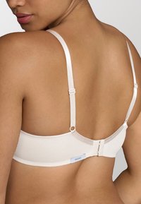 Sott bra beige chiaro con spalline regolabili, chiusura a gancio e occhiello e un'etichetta con logo vicino al lato. Texture liscia e design semplice.