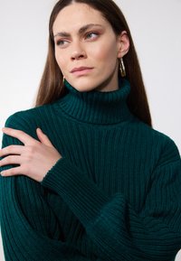 Maglione a coste di un verde scuro con colletto a coste alto, tessuto testurizzato e maniche lunghe; presenta un evidente motivo a costine e dettagli ai polsini.