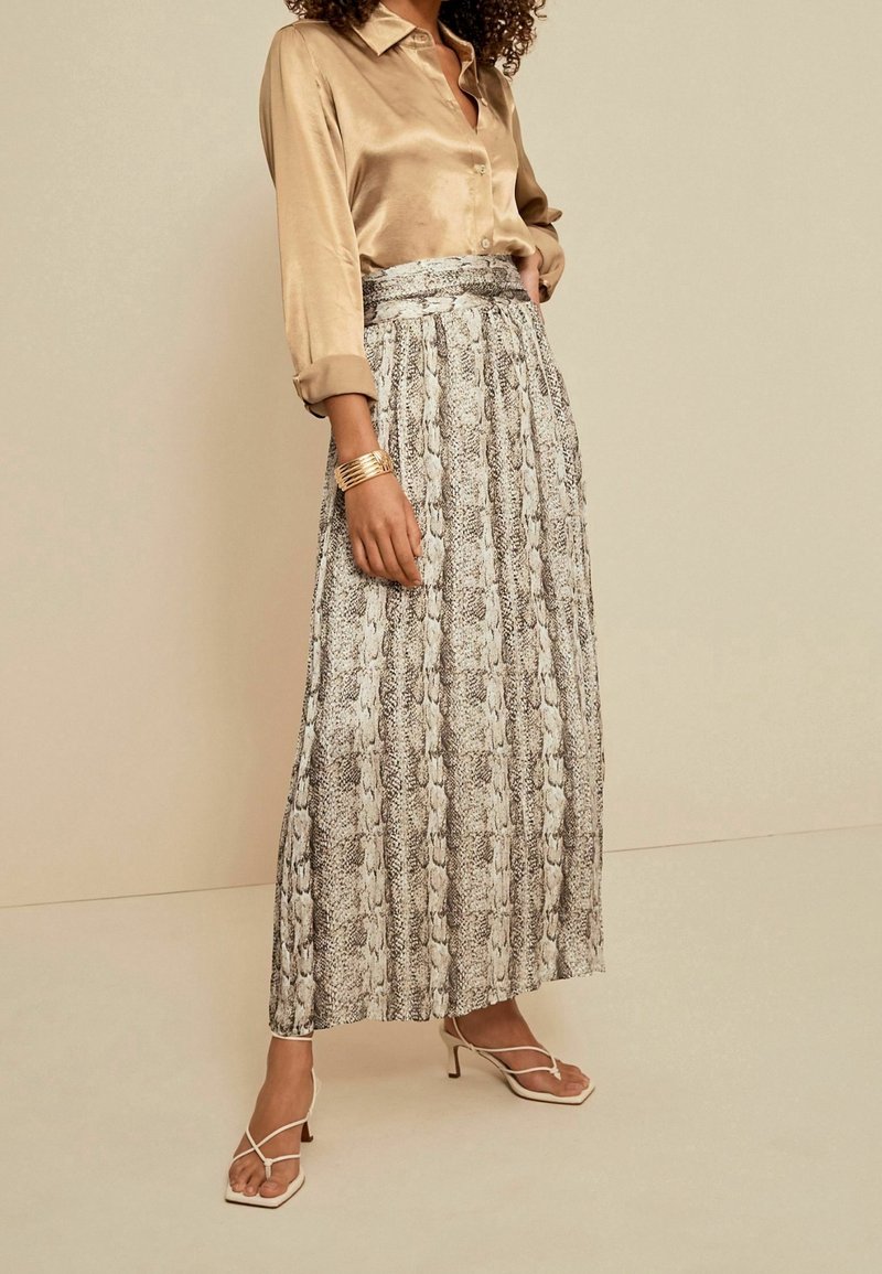 Femme portant un chemisier en satin doré, une jupe longue plissée à imprimé serpent, un bracelet manchette doré et des sandales blanches à talons et à lanières, debout contre un mur beige.