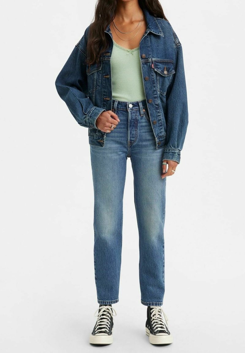 Femme portant une veste en jean bleue sur un débardeur vert clair, un jean bleu taille haute et des baskets montantes noires, debout.