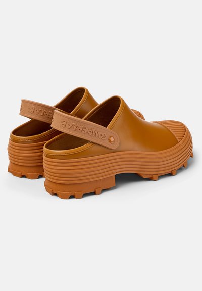 Brown rubber clogs with ridged soles and a slingback strap. Features a logo on the strap and a textured finish.