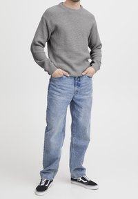 Harmaa ribbiknitti villapaita yhdistettynä vaaleansinisiin löysiin farkkuihin ja mustiin lenkkareihin, kädet taskuissa, seisoo valkoista taustaa vasten.