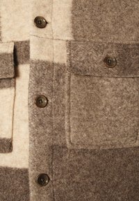 Brun og beige uldjakke med stort ternet mønster, to klaplommer og fire mørkebrune knapper foran.