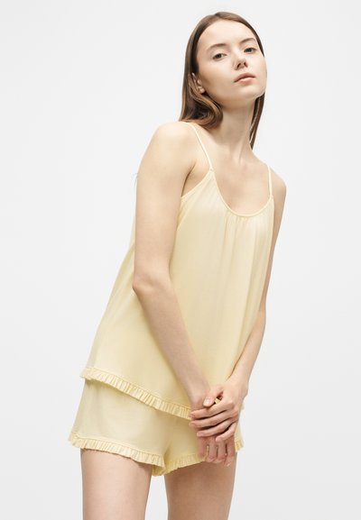Jeune femme portant un haut sans manches jaune clair et un short assorti avec un bord à volants, debout les mains jointes de manière détendue, fond neutre.