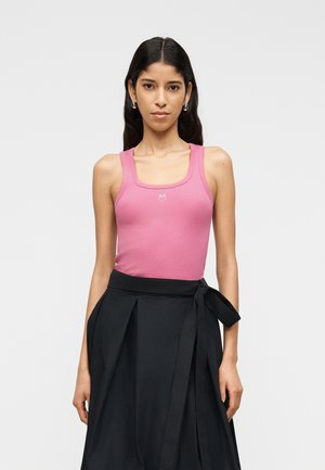 Žena s dlouhými tmavými vlasy má na sobě růžový top bez rukávů s malým emblémem a černou sukni s vysokým pasem, zavázanou v pase, na jednobarevném pozadí.