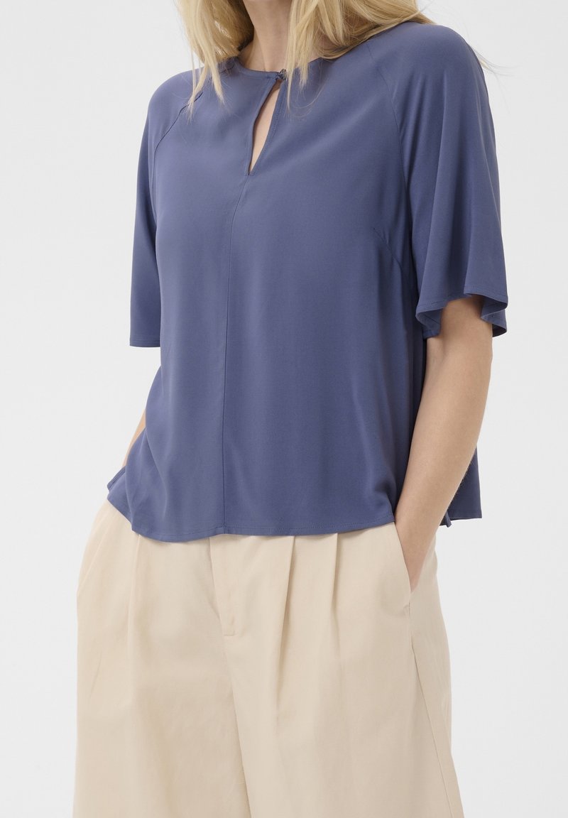 Femme portant un haut bleu ample à manches courtes avec une encoche à l'avant et un pantalon plissé beige clair avec les mains dans les poches.