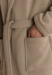 Bež ribadevoo, millel on vöö, laiad varrukad ja küljetaskud. Kangas on pehme tekstuuriga, rõhutades selle hubast disaini.